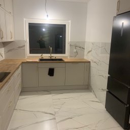 Tomasz Zegzuła - Nowoczesna kuchnia z białymi szafkami z złotymi uchwytami, drewnianym blatem, czarnym zlewem i marmurową okładziną ściany oraz podłogi, oświetlona pojedynczą żarówką.