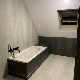 Tomasz Zegzuła - Wnętrze łazienki na poddaszu w trakcie remontu, widoczna wanna z czarną obudową, czarna armatura, surowe ściany i podłoga z płytek imitujących beton, okno dachowe.