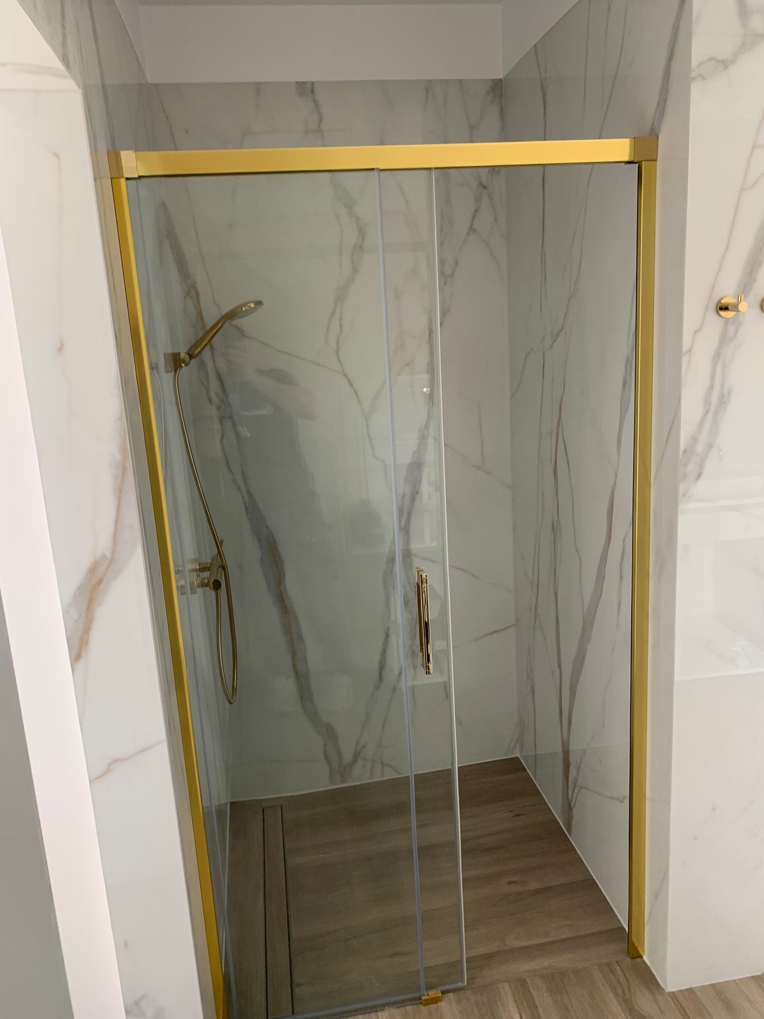 Kabina prysznicowa z przesuwnymi drzwiami o złotych profilach, marmurowe ściany i drewnopodobna podłoga w nowoczesnej łazience.