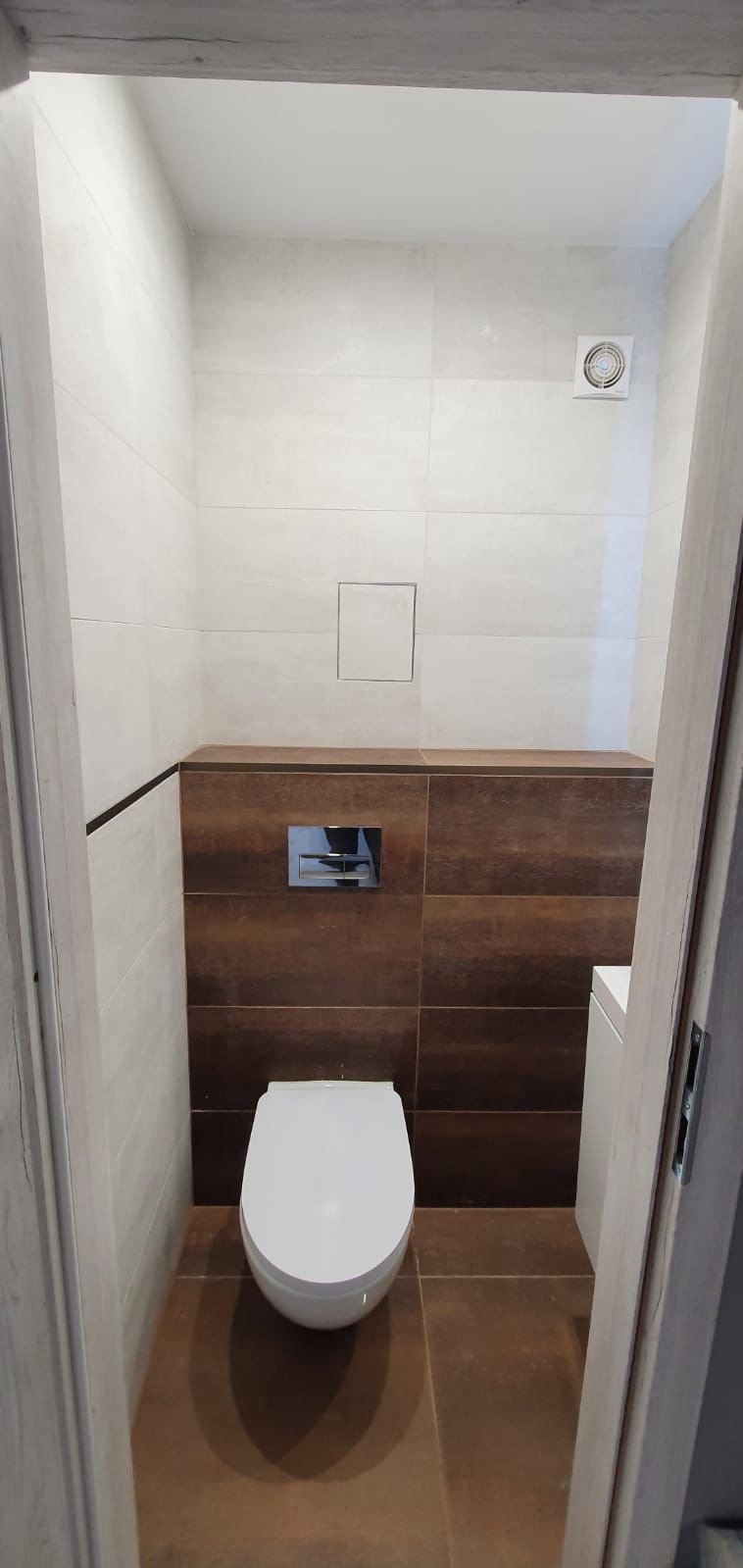 Nowoczesna toaleta podtynkowa z białym sedesem wiszącym, brązowe i beżowe płytki na ścianach, widoczna wentylacja i klapa rewizyjna.