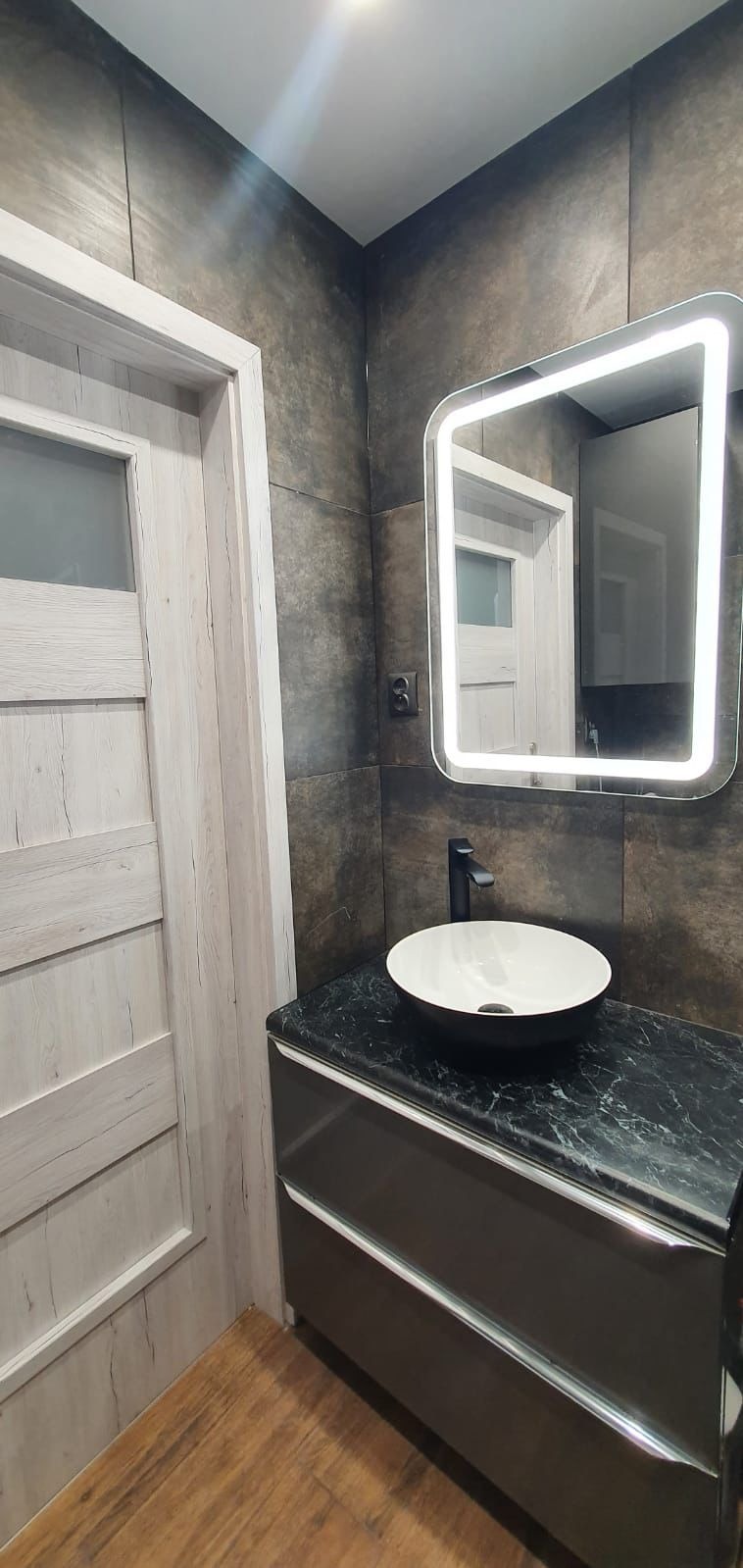 Nowoczesna łazienka z umywalką nablatową na szafce z blatem imitującym marmur, oświetlonym lustrem i grafitowymi płytkami na ścianach, widoczna drewnopodobna podłoga i drzwi z szybą.