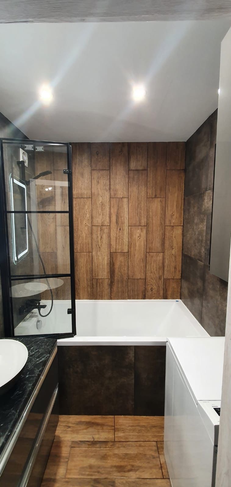 Nowoczesna łazienka z kabiną prysznicową typu walk-in z czarną ramą, białą wanną i ścianą wyłożoną płytkami imitującymi drewno, widoczne elementy umywalki z czarnym blatem.