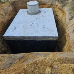 Betonowy zbiornik z pokrywą umieszczony w wykopie ziemnym, widoczne ślady stóp na górnej płycie, świeżo ułożona zaprawa wokół włazu.