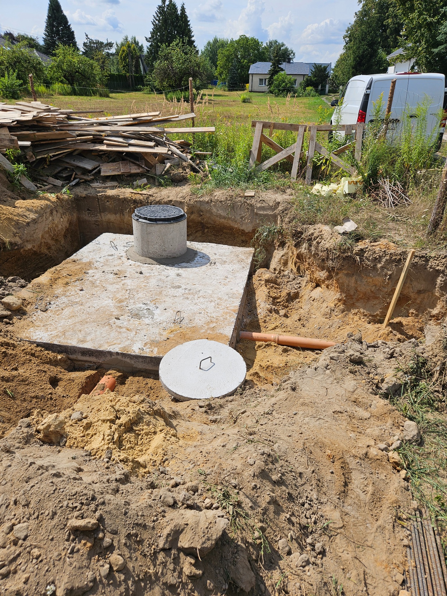 Betonowy zbiornik z pokrywą, częściowo zakopany w ziemi, z widocznymi rurami, świeżo wykopany dół, w tle deski i biały samochód dostawczy.
