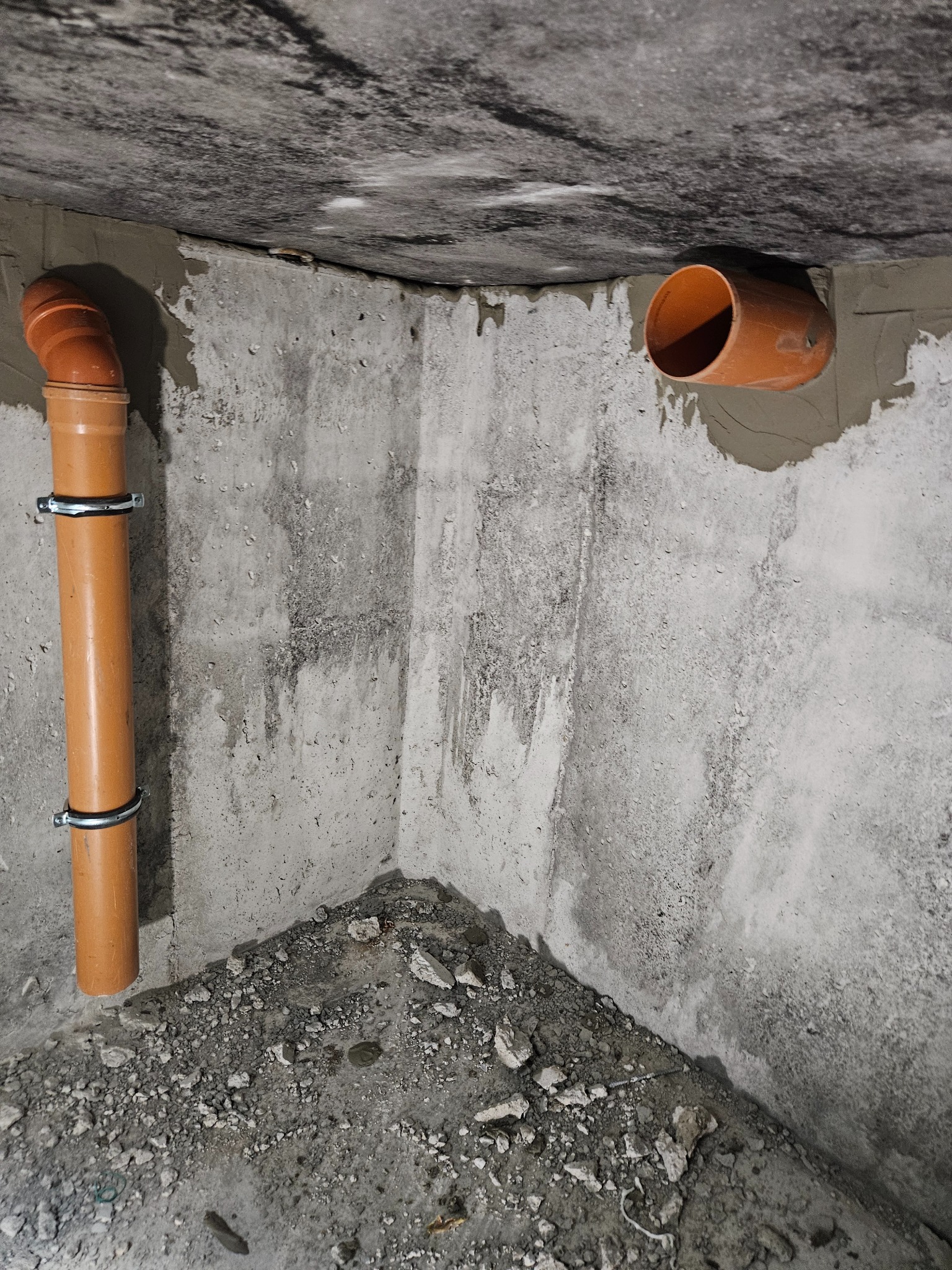 Pomarańczowa rura kanalizacyjna pionowa przymocowana do surowej, betonowej ściany z widocznymi śladami po zaprawie i gruzem na posadzce. Druga rura pozioma wystaje ze ściany po prawej stronie.
