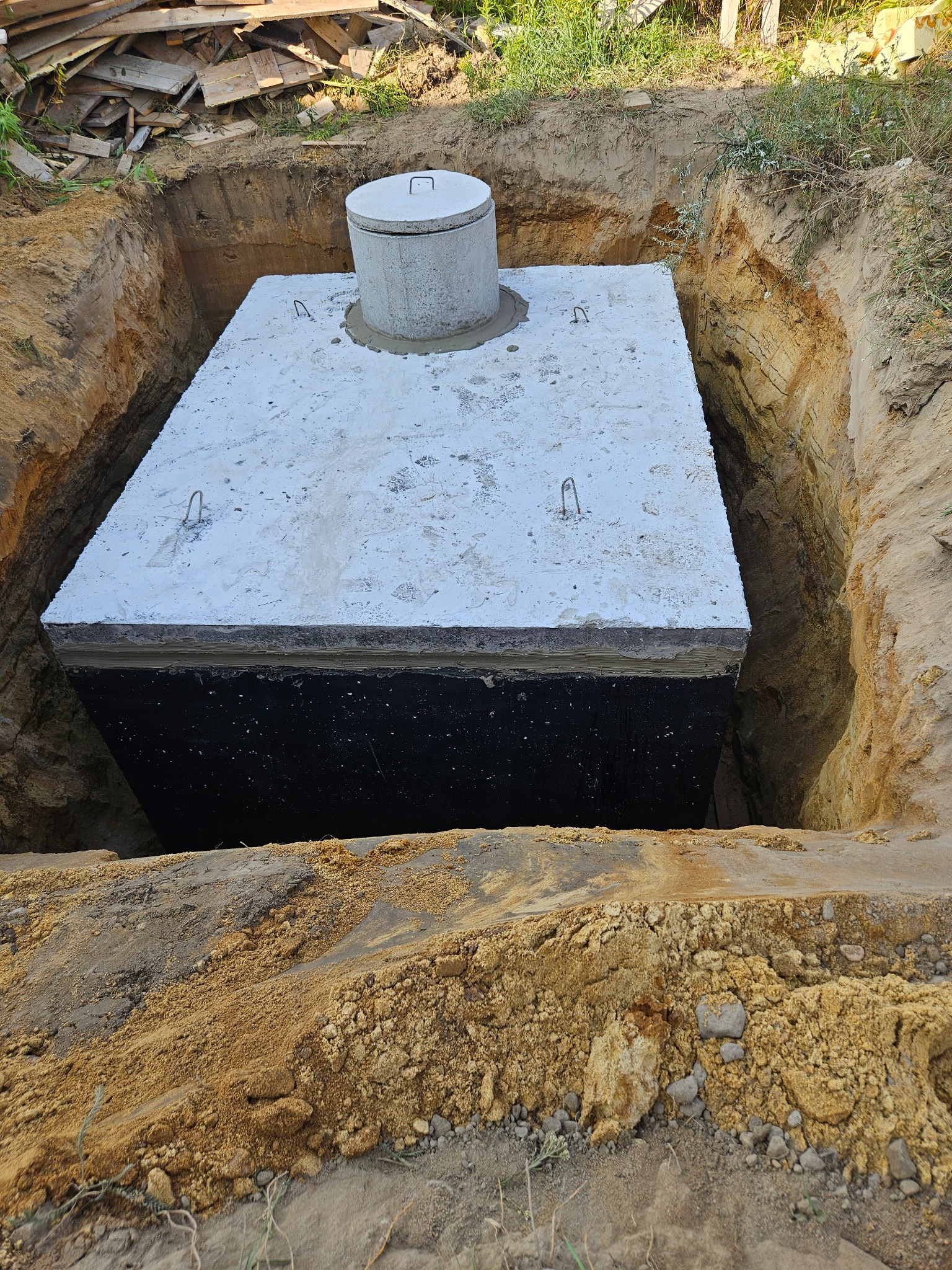 Betonowy zbiornik z pokrywą umieszczony w wykopie ziemnym, widoczne ślady stóp na górnej płycie, świeżo ułożona zaprawa wokół włazu.
