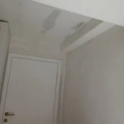 Ściana i sufit w trakcie szpachlowania, widoczne nierówności i nałożona gładź, białe drzwi z metalową klamką po lewej stronie.