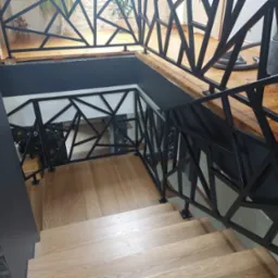 Nowoczesne schody drewniane z czarną, geometryczną balustradą metalową. Widoczne schody z jasnego drewna i fragment podłogi na górze. W tle odkurzacz przemysłowy.