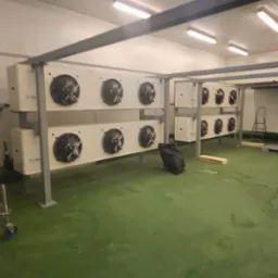 Wnętrze hali z zamontowanymi agregatami chłodniczymi, widoczne stalowe konstrukcje wsporcze, drabina i rusztowanie na kółkach.