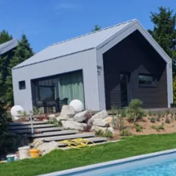 Nowoczesny, dwukolorowy dom z szarymi oknami i drzwiami przesuwnymi, widok z perspektywy basenu, z widocznymi schodami prowadzącymi do wejścia, elementami dekoracyjnymi i narzędziami malarskimi.