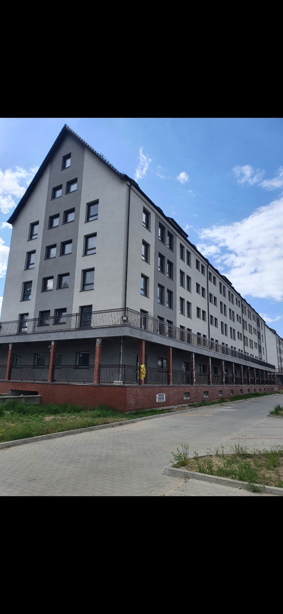 Szary budynek wielorodzinny z balkonami i oknami, widok z dołu na tle błękitnego nieba z chmurami.