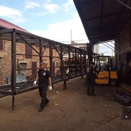 Stal Company - Montaż stalowej konstrukcji pomostu łączącego budynki, widoczni pracownicy w roboczych ubraniach i wózki widłowe.