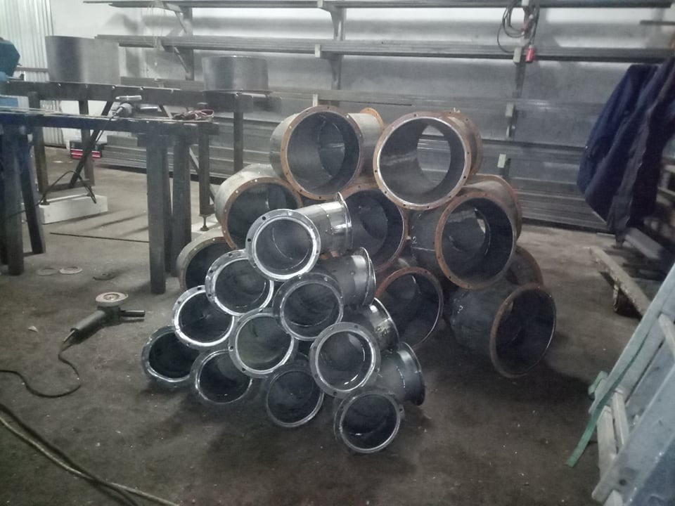 Stalowe rury i kolanka z kołnierzami ułożone na podłodze warsztatu, szlifierka kątowa obok, metalowe regały w tle.