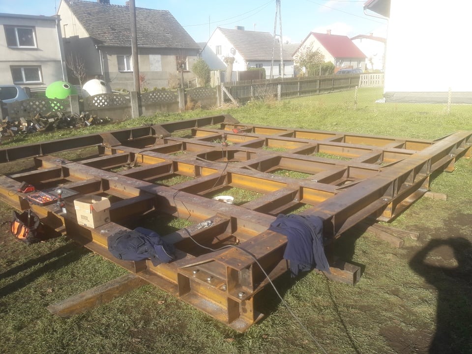 Szkielet stalowej konstrukcji na trawie, widoczne narzędzia i karton, w tle budynki mieszkalne.