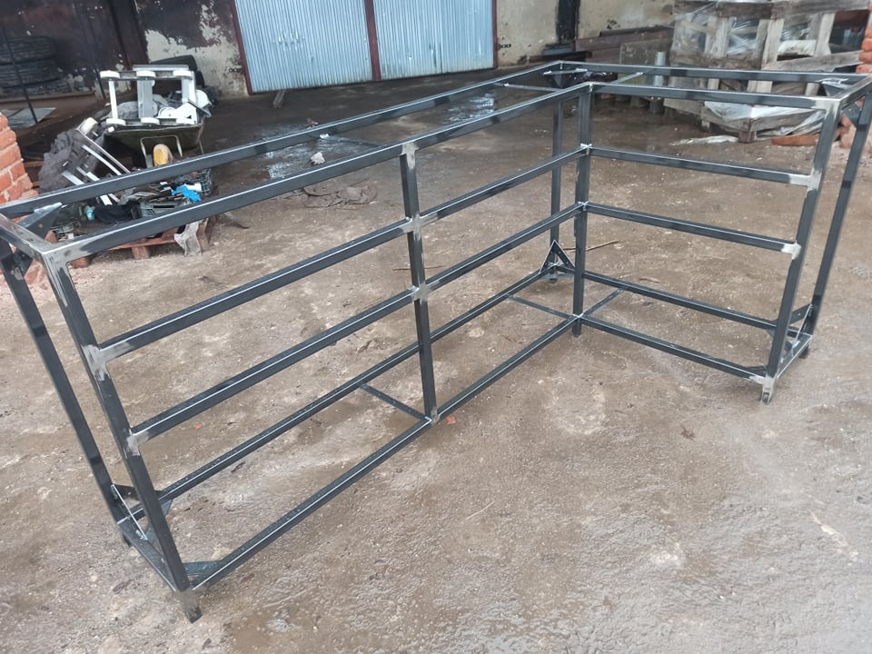 Metalowa konstrukcja szkieletowa w kształcie litery L, spawana z profili, z widocznymi śladami spawania, na tle betonowego podłoża i elementów warsztatu.