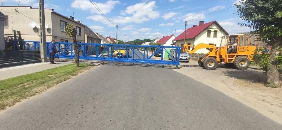 Transport niebieskiej konstrukcji stalowej przy użyciu żółtej ładowarki na tle wiejskich domów i błękitnego nieba.