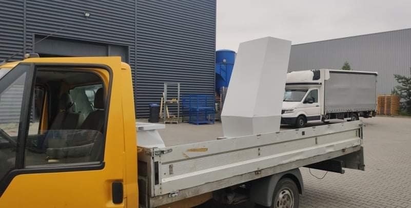 Żółty samochód dostawczy z białą, metalową konstrukcją w kształcie komina na pace, na tle szarego budynku przemysłowego i białej furgonetki