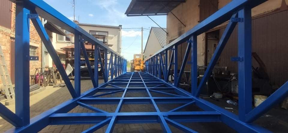 Pomarańczowy wózek widłowy w tle, widoczny przez konstrukcję stalową pomalowaną na niebiesko, z perspektywy podłoża.