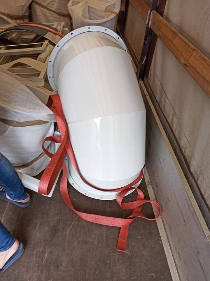 Biały, metalowy element konstrukcyjny w kształcie cylindra z kołnierzami, zabezpieczony czerwoną taśmą transportową w przestrzeni ładunkowej, widoczne fragmenty innych elementów i osoba w klapkach.