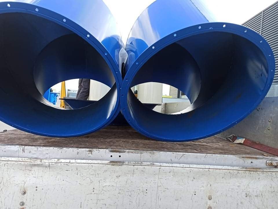 Dwa duże, niebieskie, cylindryczne elementy metalowe z kołnierzami na krawędziach, leżące na skrzyni ładunkowej pojazdu. Wnętrze elementów jest ciemniejsze, widoczne ślady spawania. W tle budynek...