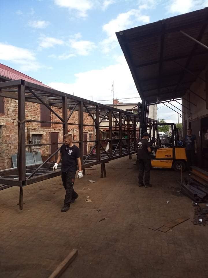 Montaż stalowej konstrukcji pomostu łączącego budynki, widoczni pracownicy w roboczych ubraniach i wózki widłowe.