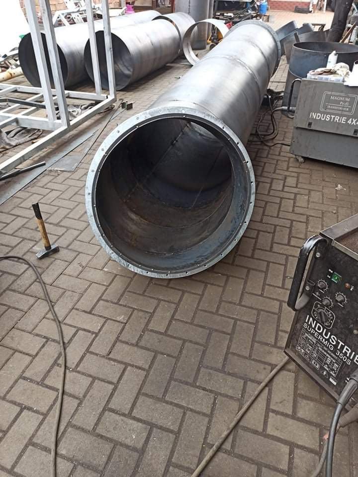 Duży element stalowy rury z widocznymi spawami na krawędziach, leżący na brukowanej powierzchni, obok konstrukcji szkieletowej i urządzeń do obróbki metali.