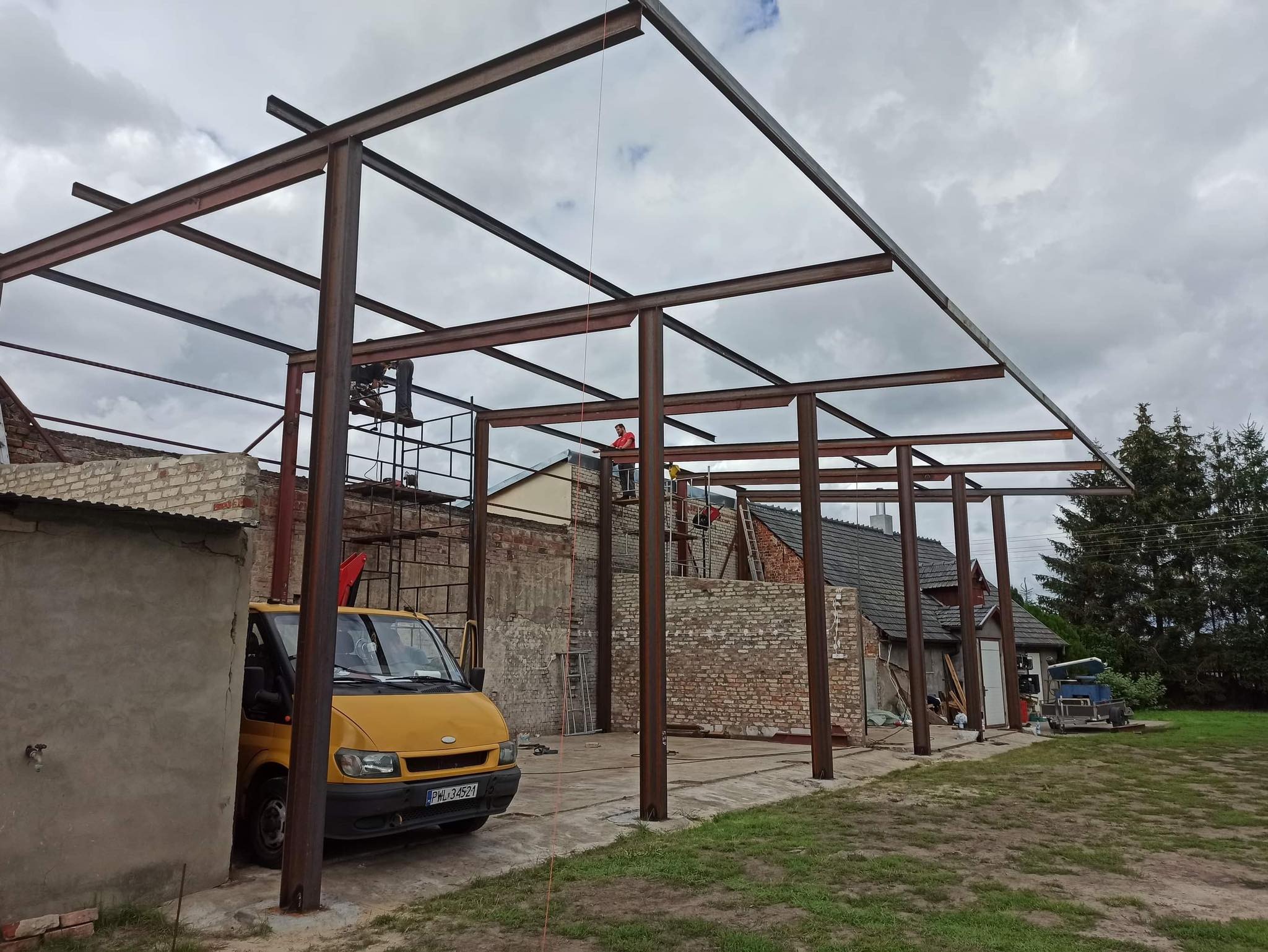 Konstrukcja stalowa hali w trakcie budowy, widoczni robotnicy na rusztowaniach, żółty bus zaparkowany obok, pochmurne niebo w tle.