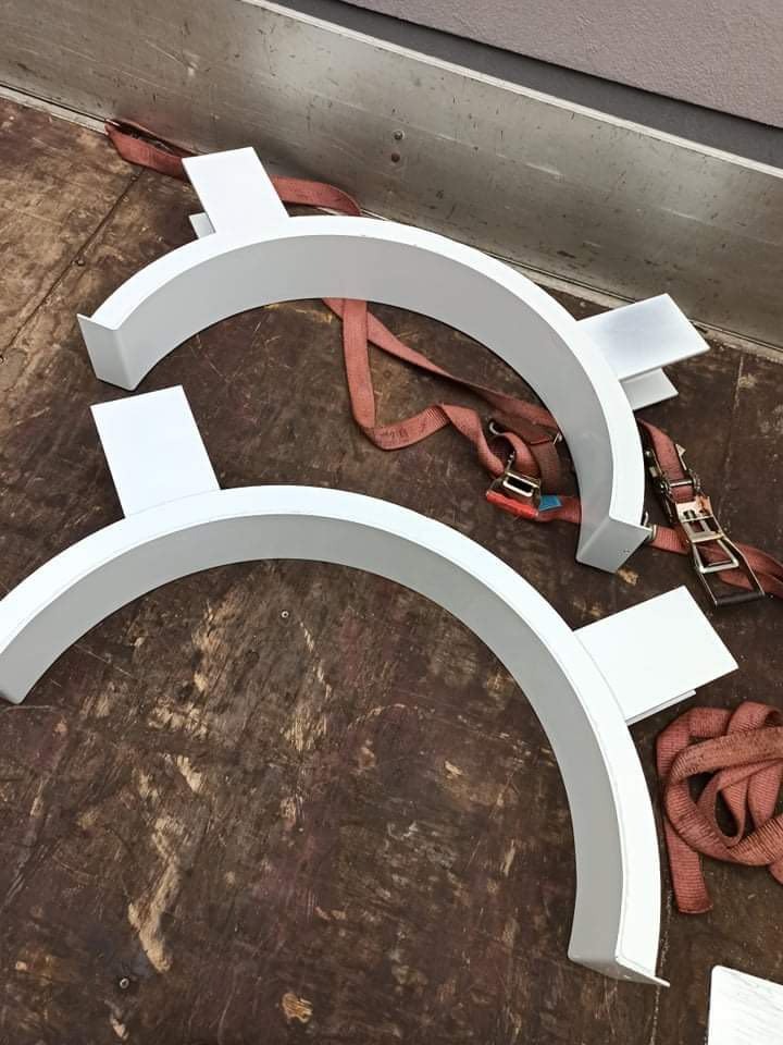 Dwa białe, półokrągłe elementy konstrukcyjne z metalu, z przyspawanymi płaskownikami, zabezpieczone pasami transportowymi na brudnej powierzchni.