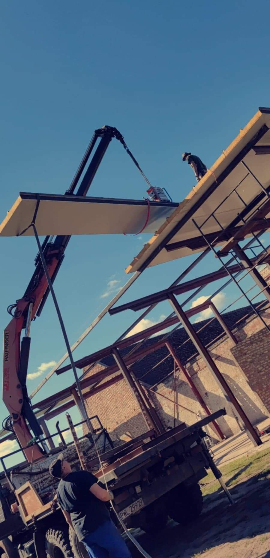 Montaż paneli dachowych na konstrukcji stalowej hali za pomocą dźwigu, widoczny pracownik na dachu oraz fragment starego budynku w tle.