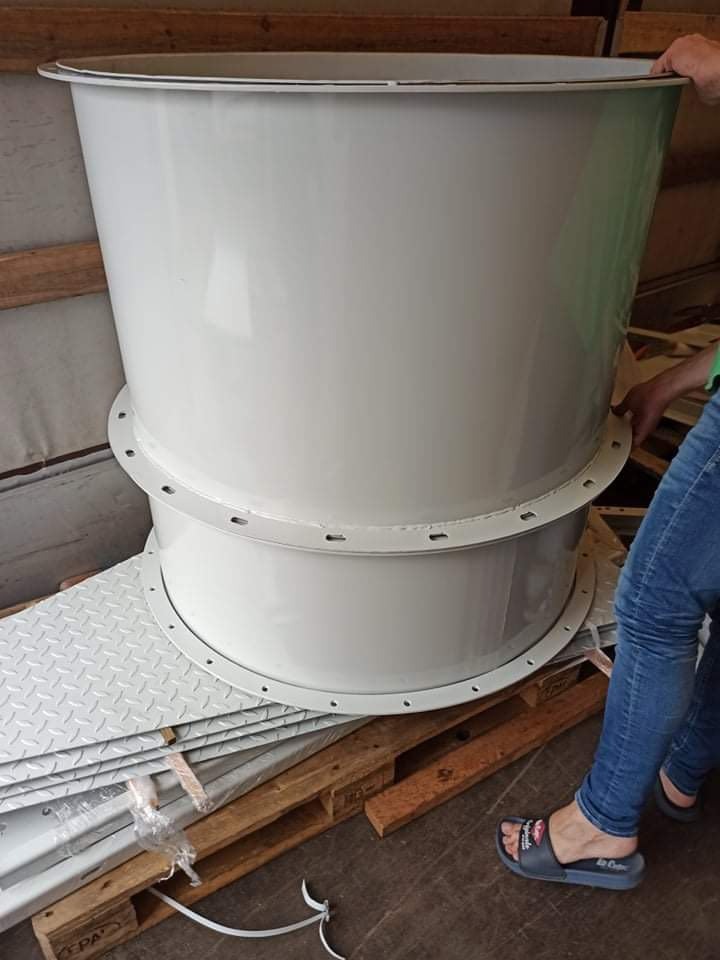 Dwa segmenty białej, metalowej rury wentylacyjnej z kołnierzami, leżące na palecie. Widoczne nogi osoby w dżinsach i klapkach.