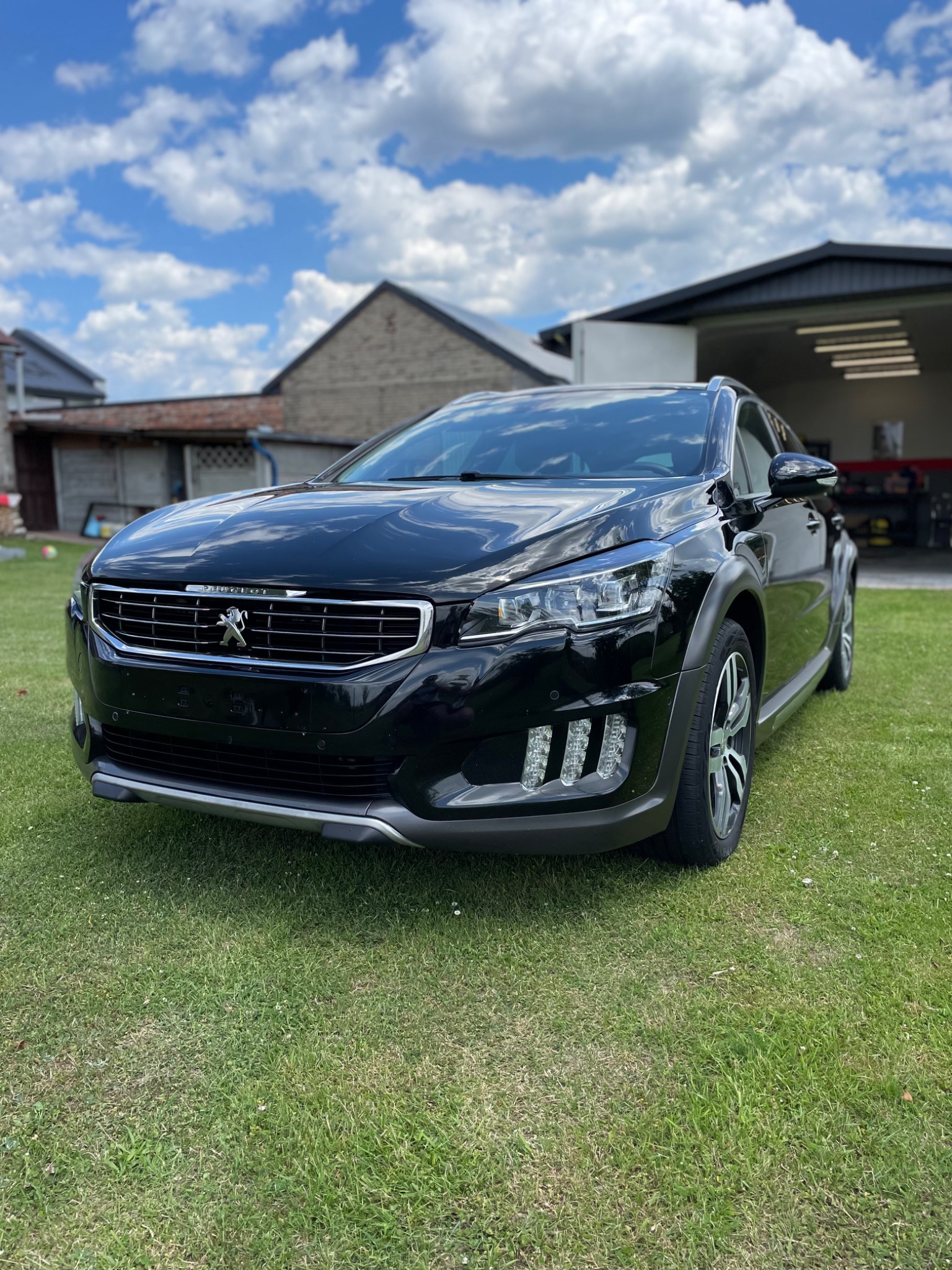Błyszczący, czarny Peugeot 508 RXH stoi na trawniku przed garażem w słoneczny dzień, z widocznymi detalami grilla i reflektorów.