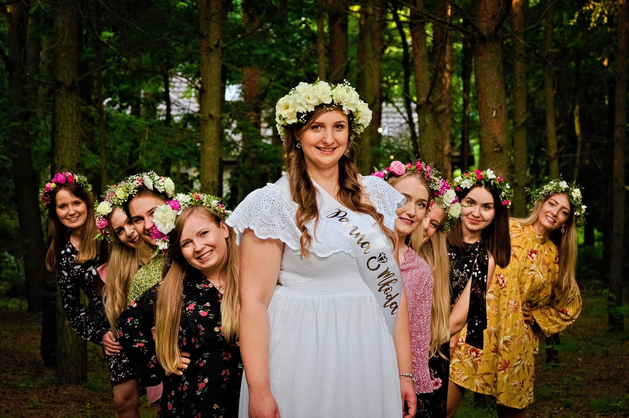 Panna młoda w białej sukience i wianku na głowie, otoczona druhnami w wiankach, pozuje na tle leśnego krajobrazu.