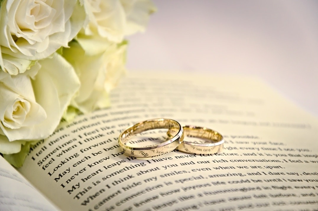 Dwa złote obrączki ślubne leżące na otwartej książce z bukietem białych róż w tle.