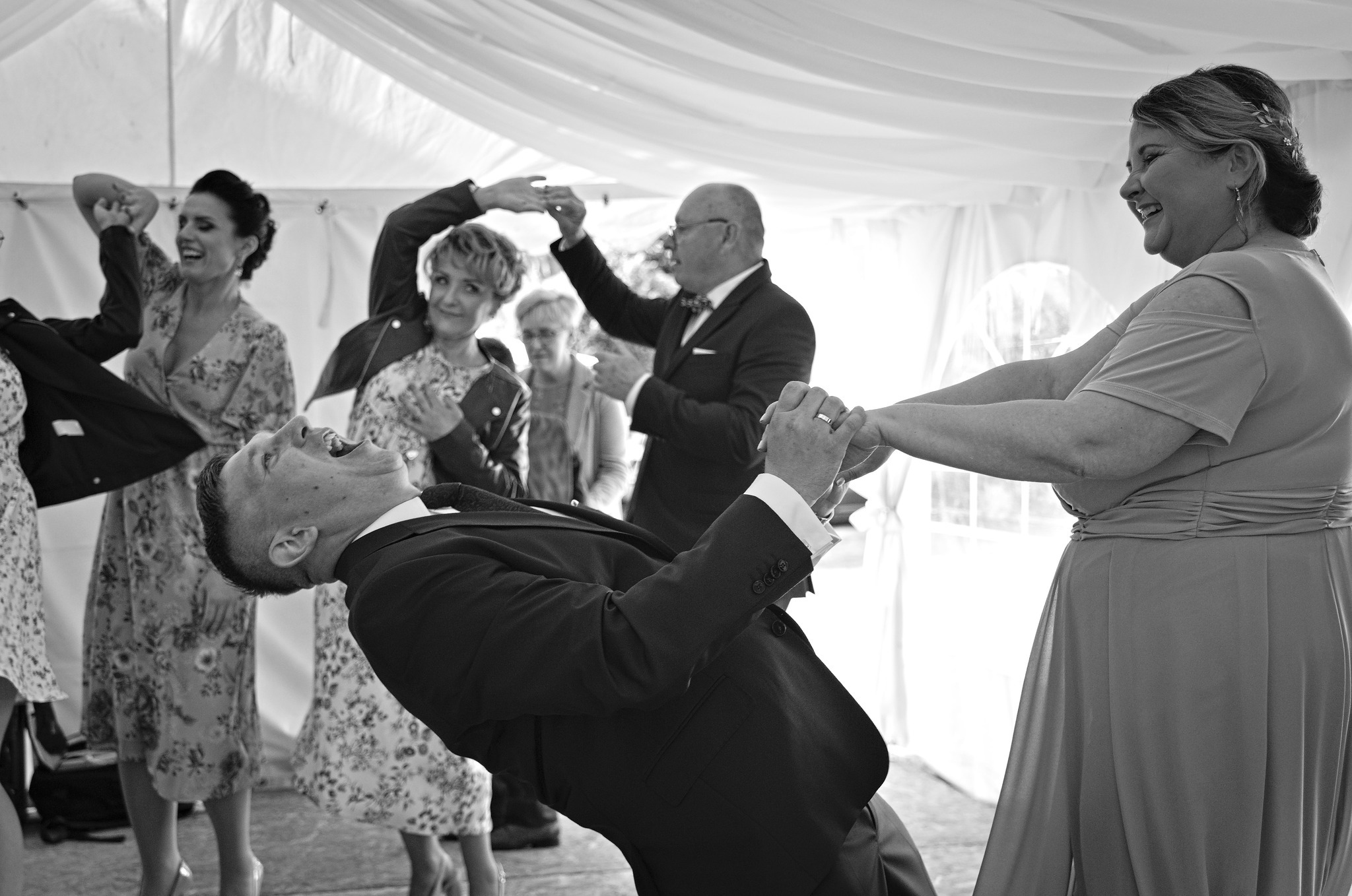 Czarno-białe zdjęcie weselne: Pan młody w czarnym garniturze odchyla się do tyłu z otwartymi ustami podczas oczepin, trzymany za ręce przez uśmiechniętą kobietę w sukience, w tle goście weselni...