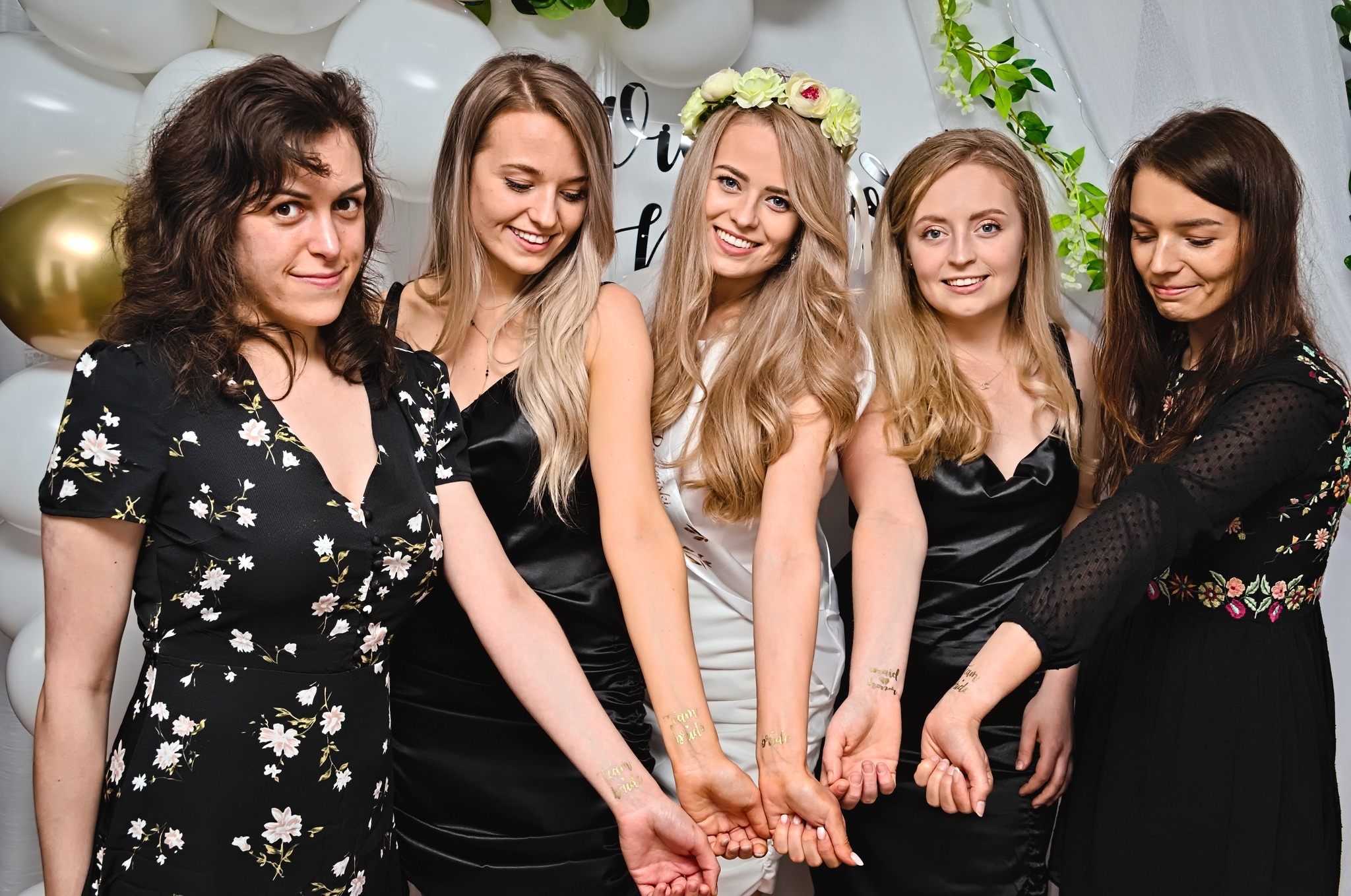 Pięć kobiet w sukienkach, z tymczasowymi tatuażami 'Team Bride' na nadgarstkach, pozuje na tle białych balonów i napisu 'cheers', panna młoda w wianku.