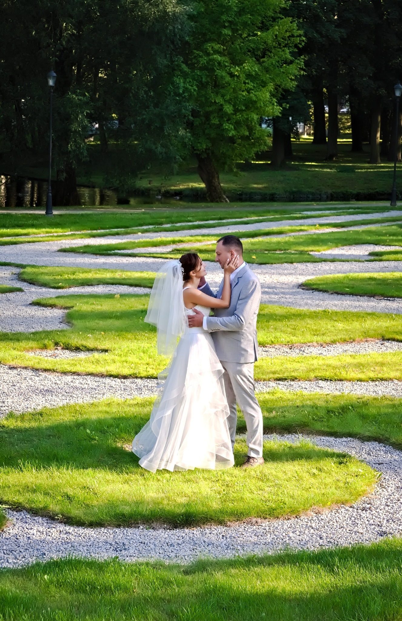 Para młoda w objęciach na tle parku z geometrycznym wzorem trawników i żwirowych ścieżek, panna młoda w zwiewnej sukni z welonem, pan młody w jasnoszarym garniturze.