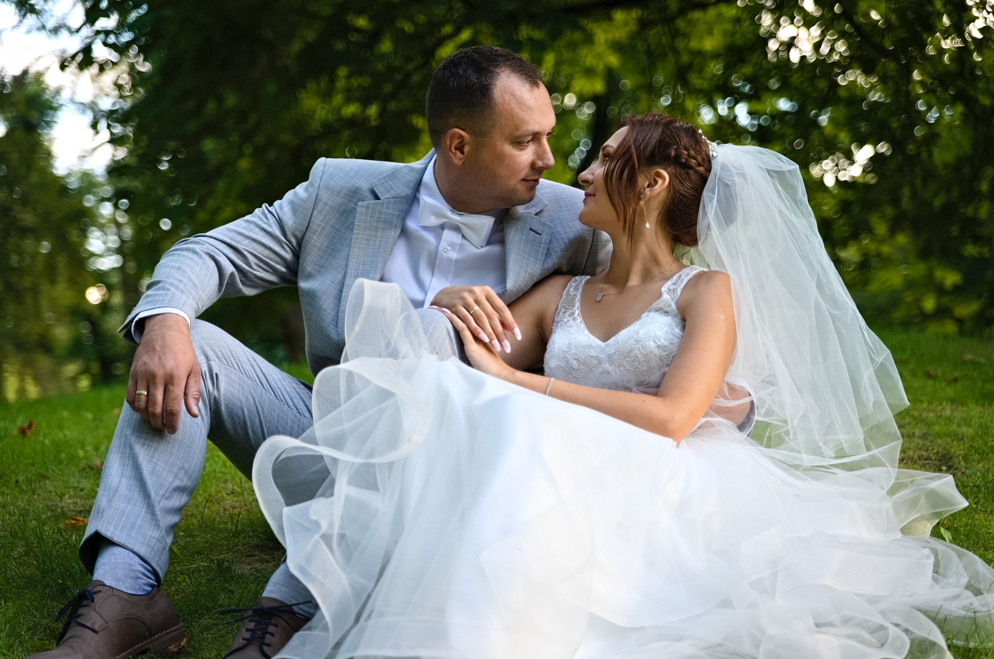 Para młoda siedzi na trawie, panna młoda w białej sukni z koronkową górą i welonem, pan młody w szarej marynarce i muszce, patrzą na siebie w zamyśleniu, w tle zieleń drzew.