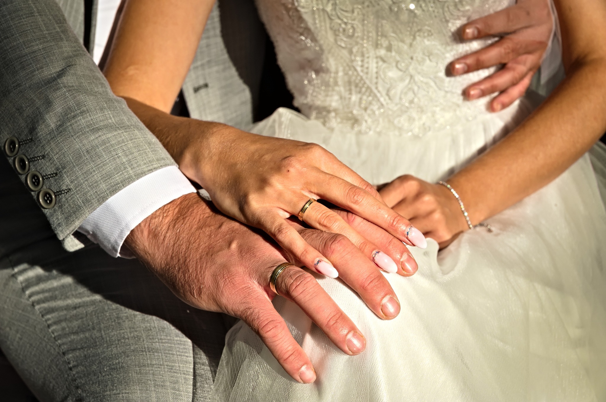 Dłonie pary młodej z obrączkami na tle sukni ślubnej i marynarki w kratę.