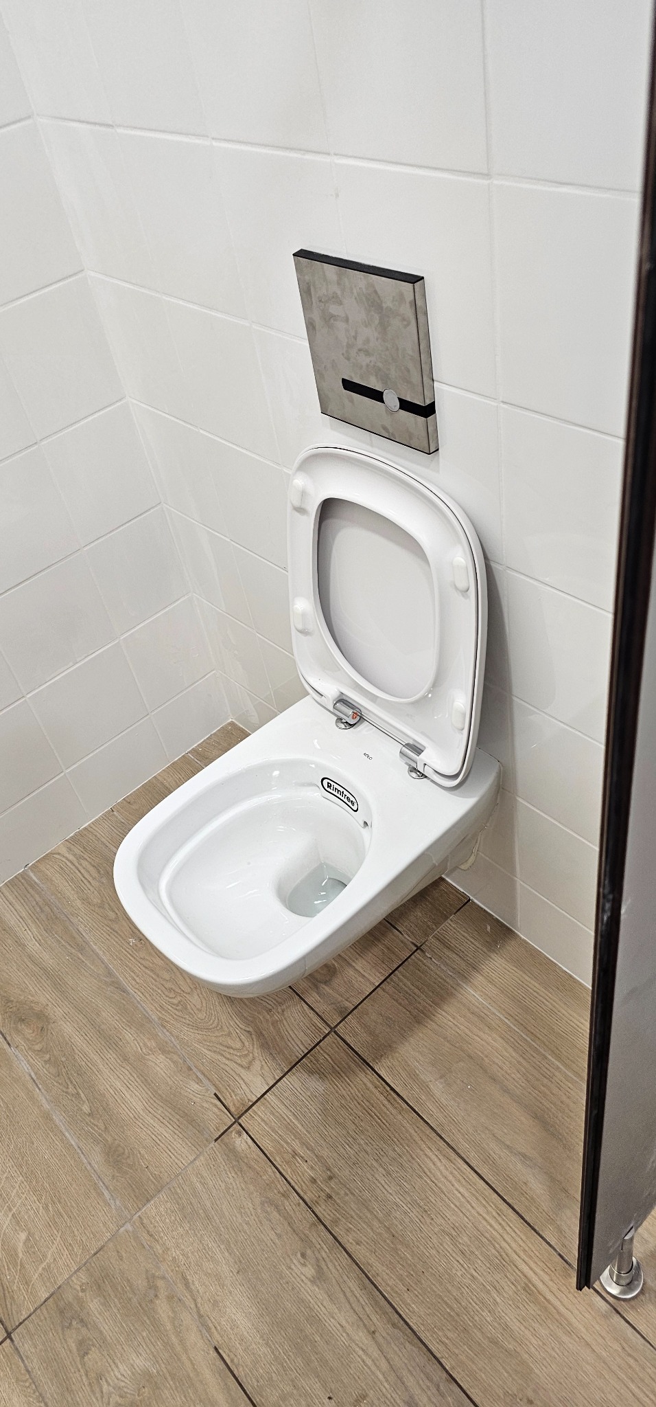 Wisząca toaleta marki Koło Rimfree z otwartą deską, zamontowana na białych, kwadratowych płytkach ściennych i drewnopodobnych płytkach podłogowych, z widocznym szarym panelem spłukującym.