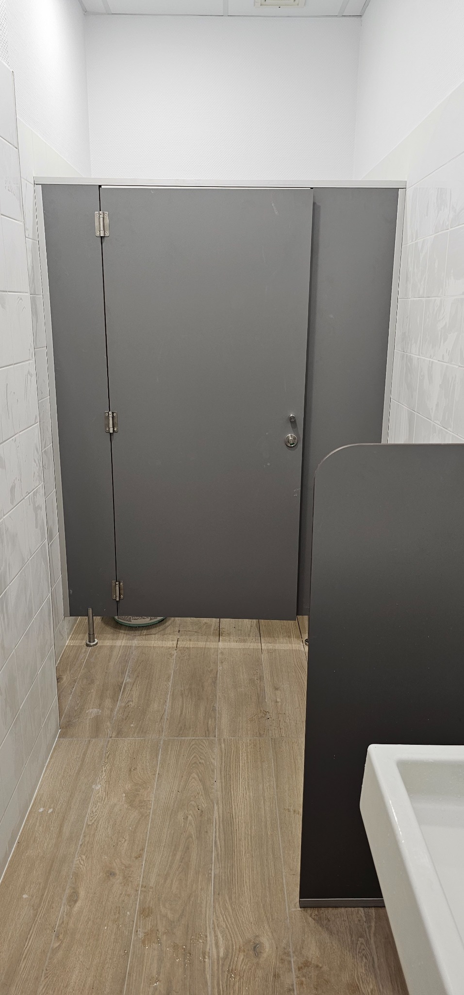 Szara kabina toaletowa z metalowymi zawiasami i zamkiem w publicznej toalecie, widoczne płytki podłogowe imitujące drewno i białe płytki na ścianach.