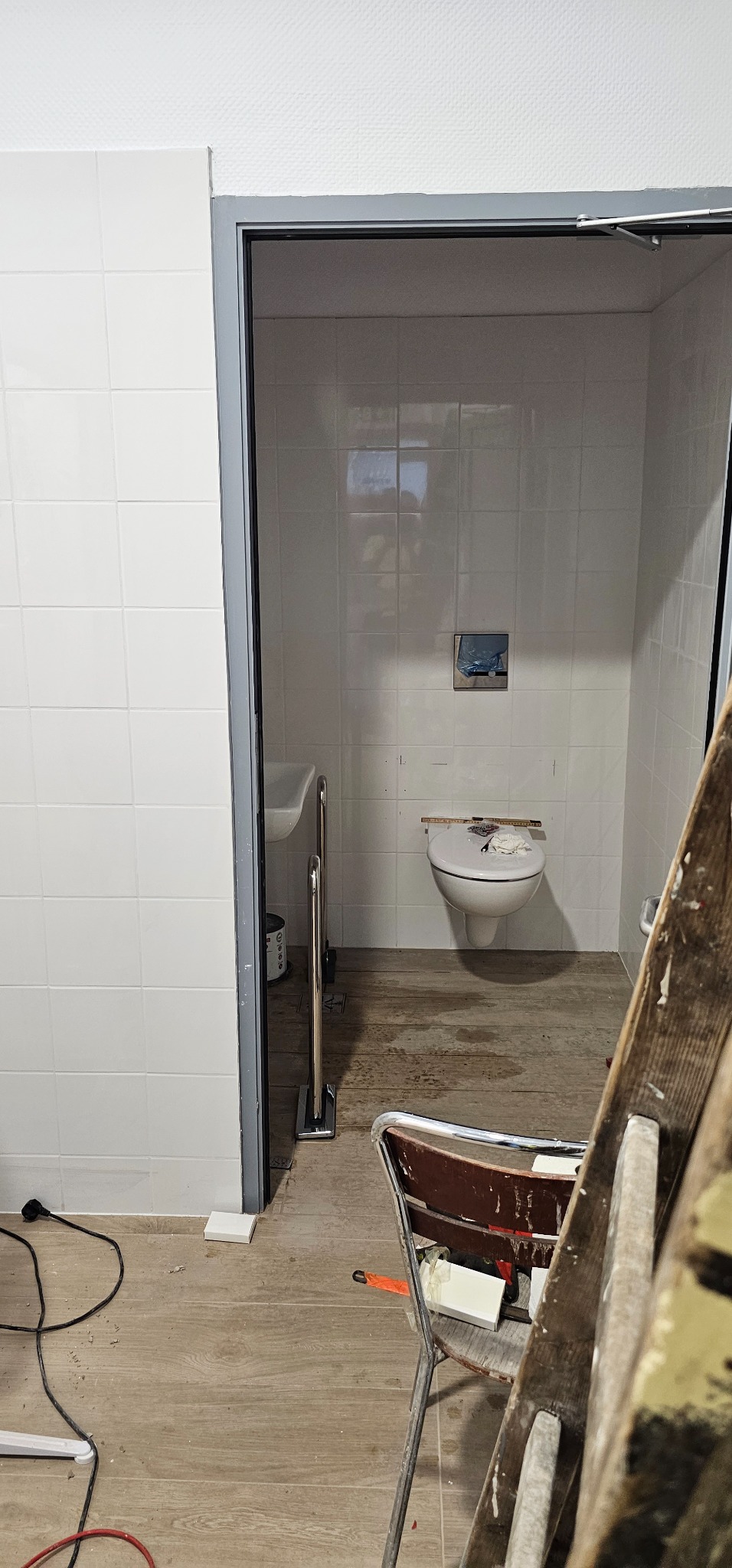 Wnętrze łazienki w trakcie prac wykończeniowych, widoczne białe płytki na ścianach, umywalka, toaleta z przykryciem oraz drabina i krzesło malarskie z narzędziami.