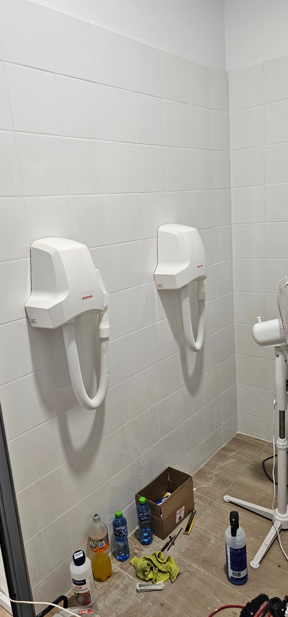 Dwa białe suszarki do rąk marki Starmix zamontowane na białych kafelkach w łazience, na podłodze butelki z napojami, karton, narzędzia i ścierka, widoczny fragment białego wentylatora.