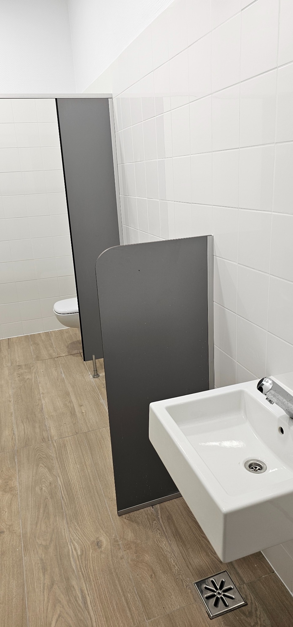 Nowoczesna łazienka publiczna z białymi płytkami, drewnopodobną podłogą, kabinami z szarego laminatu i białą umywalką z metalowym kranem