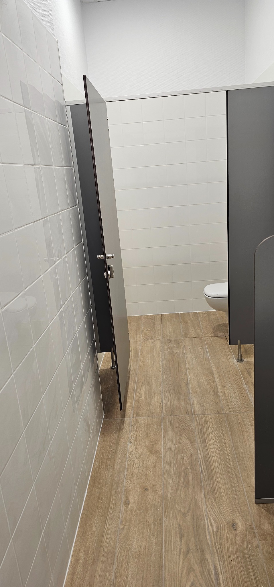 Wnętrze toalety publicznej z kabinami z szarymi drzwiami, jasnymi płytkami na ścianach i podłogą imitującą drewno, widoczny fragment toalety.