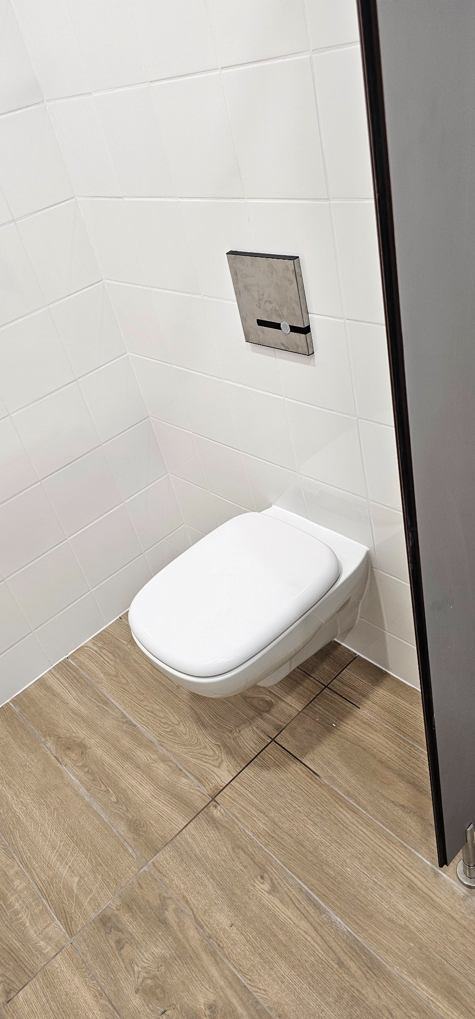 Wisząca toaleta w nowoczesnej łazience z białymi płytkami ściennymi i podłogą imitującą drewno, widoczny panel sterujący spłukiwaniem o fakturze betonu.