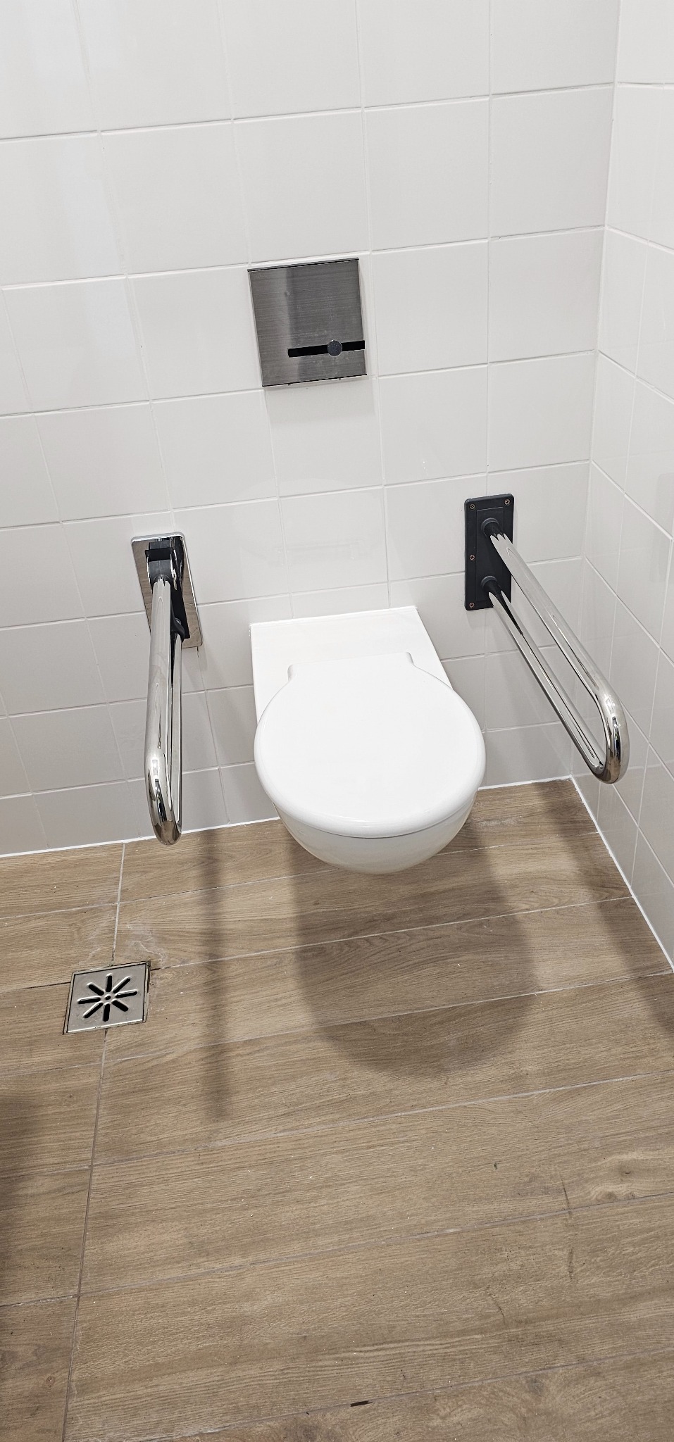 Biała toaleta wisząca z uchwytami dla niepełnosprawnych na tle białych płytek i drewnopodobnej podłogi z odpływem liniowym.