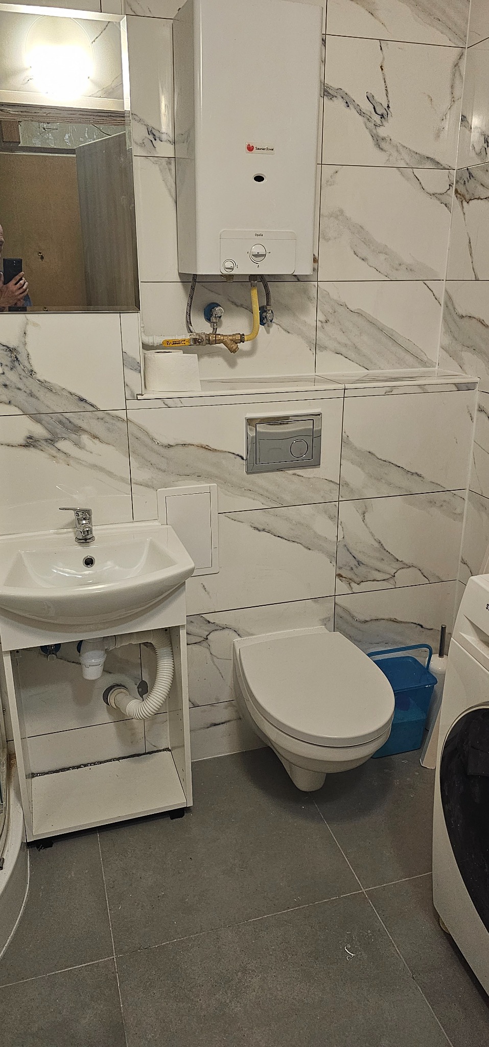 Wnętrze łazienki z białymi marmurkowymi płytkami, wiszącą toaletą, umywalką z szafką, bojlerem Saunier Duval i fragmentem pralki.