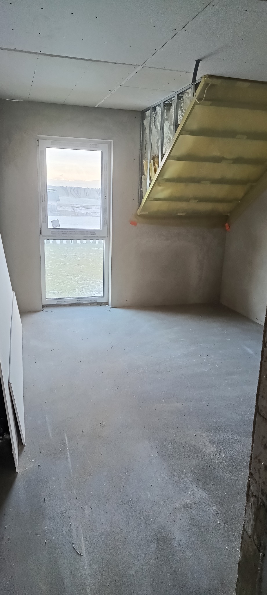 Wnętrze w stanie surowym: ściany z szarym tynkiem, okno z widokiem na zewnątrz, fragment konstrukcji poddasza z izolacją. Widoczne ślady po pracach budowlanych.