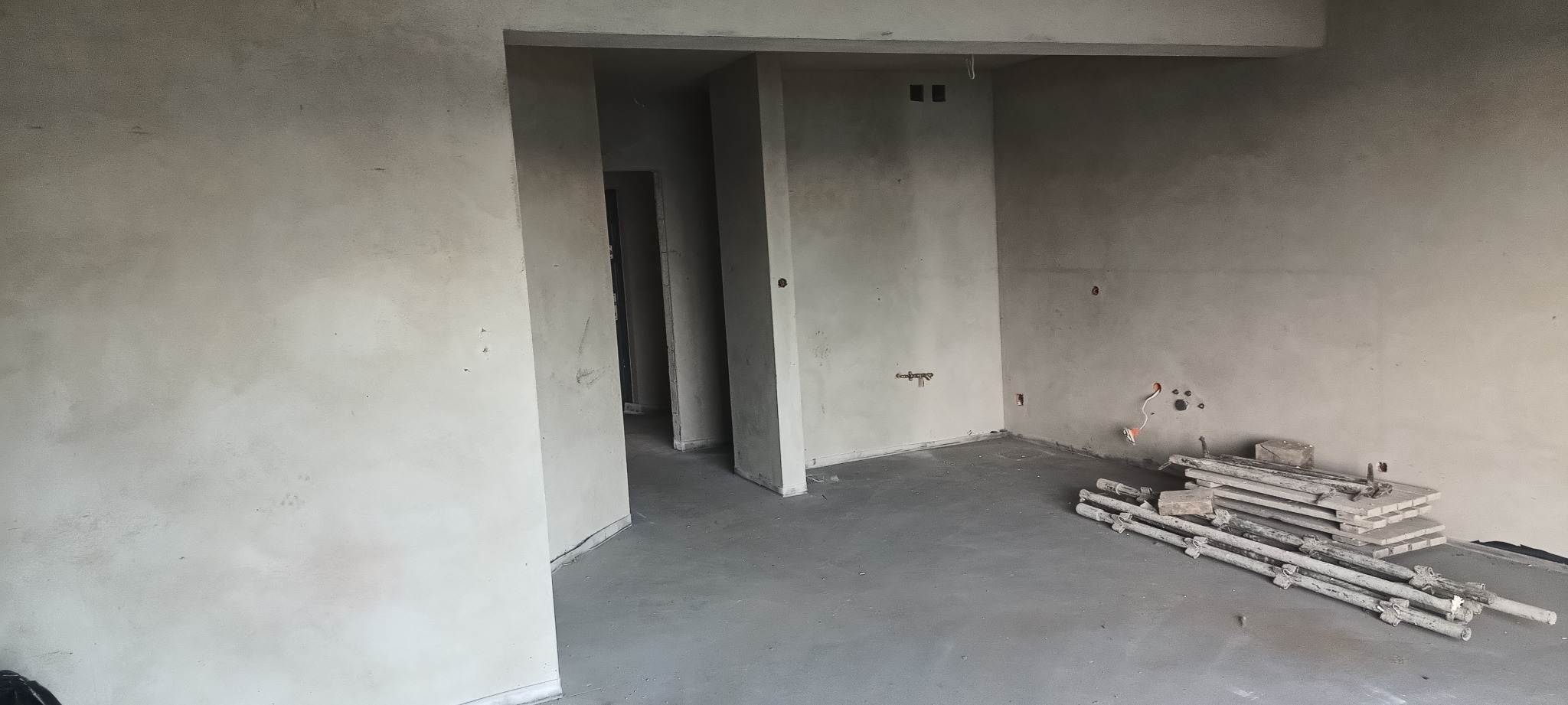 Wnętrze po tynkowaniu maszynowym: gładkie, szare ściany, widoczne instalacje elektryczne i hydrauliczne, oraz stos drewnianych podpór na podłodze.