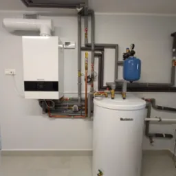 Biały kondensacyjny kocioł gazowy marki Buderus z podłączonym zasobnikiem ciepłej wody i niebieskim naczyniem wzbiorczym, widoczne rury instalacji grzewczej z izolacją termiczną.